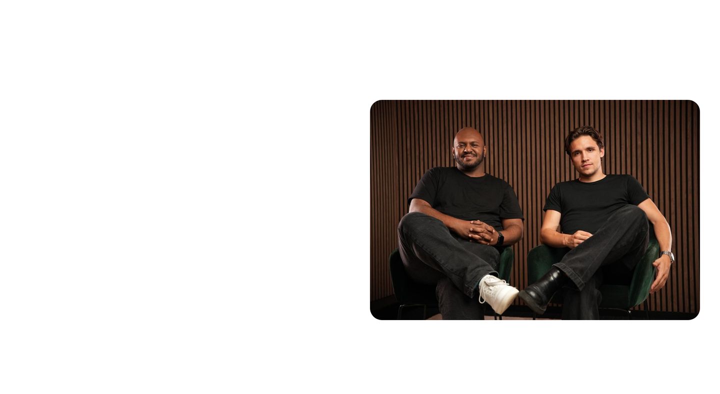 Expansive white space gives way to an offset portrait frame of two founders seated against a warm wood-slatted wall.